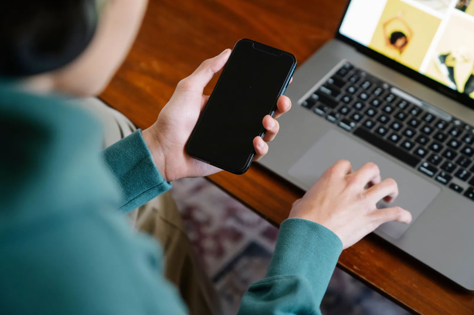 Mãos seguram um celular e mexem no touchpad de um notebook sobre uma mesa de madeira