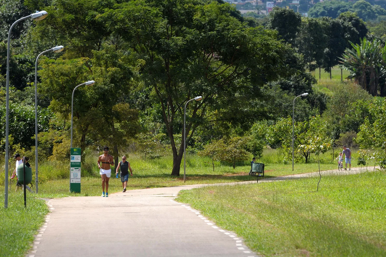 Pessoas correm e caminham em pista de concreto entre árvores e postes de luz no Parque Ecológico de Águas Claras
