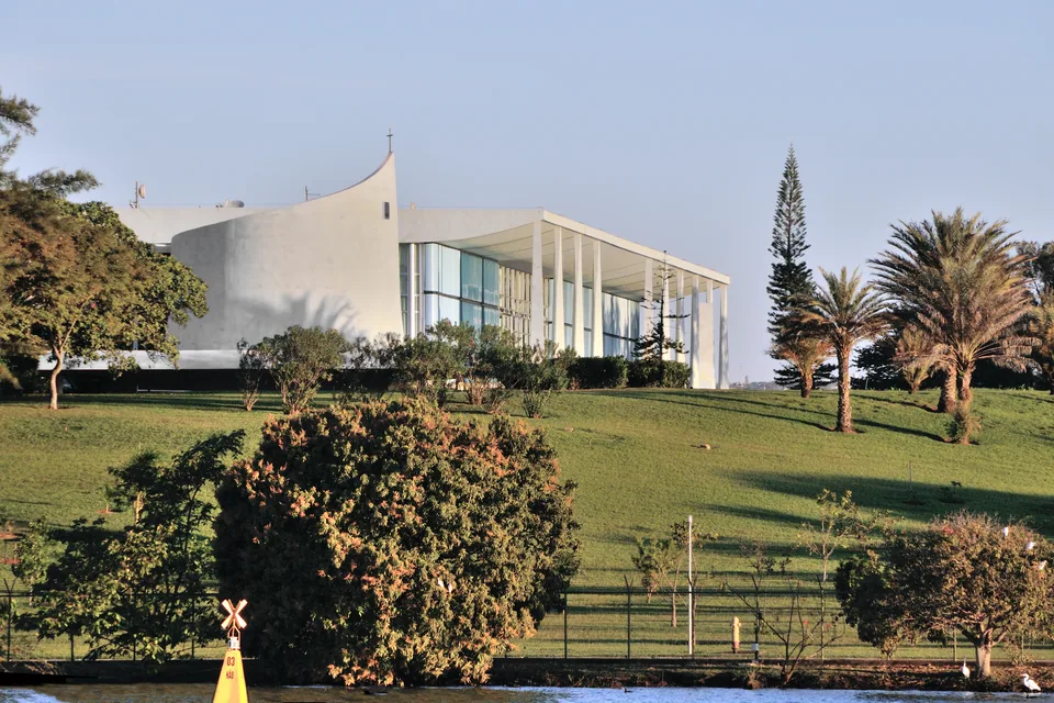 Palácio da Alvorada visto da margem do Lago Paranoá, com gramado verde, palmeiras e céu azul