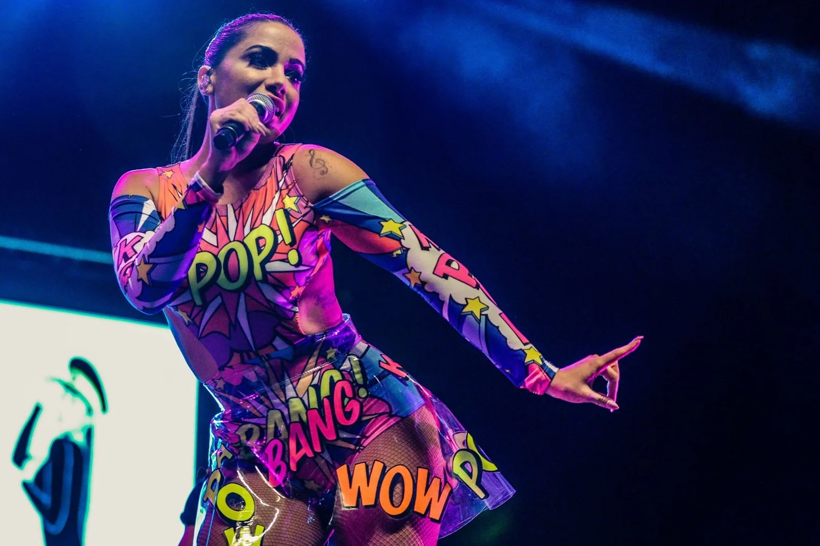 Anitta cantando no palco, com microfone na mão e figurino colorido