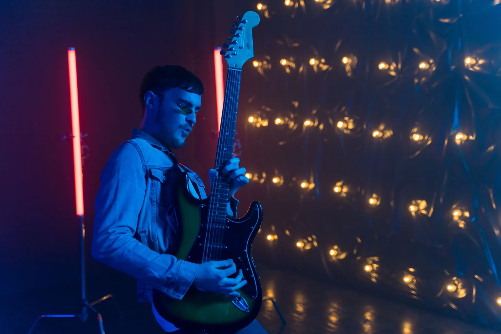 Guitarrista de jaqueta jeans toca guitarra elétrica sob luzes de neon e lâmpadas amarelas