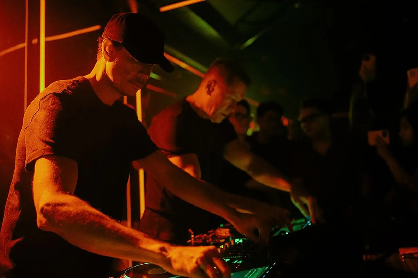 DJs mixando em cabine de balada com iluminação de neon laranja e verde