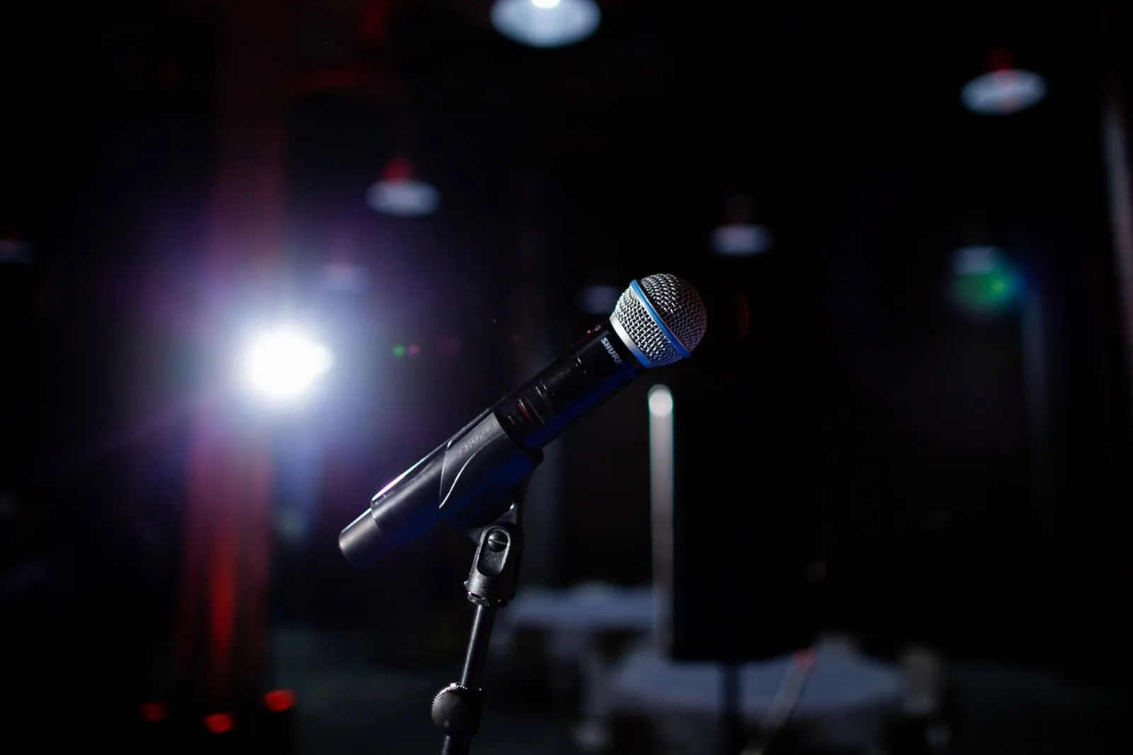 Microfone em pedestal num palco escuro, com um refletor aceso ao fundo