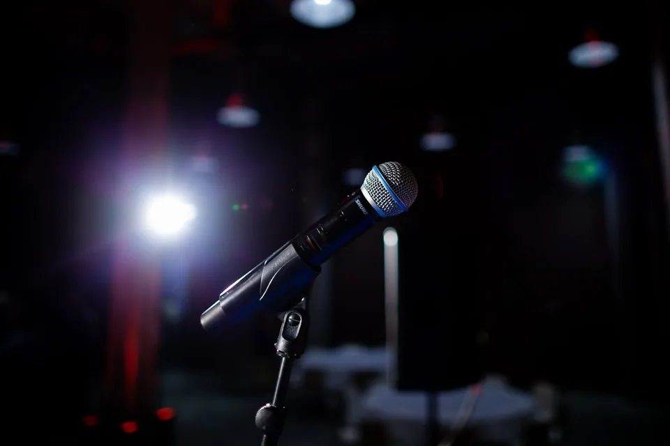 Microfone em pedestal num palco escuro, com um refletor aceso ao fundo
