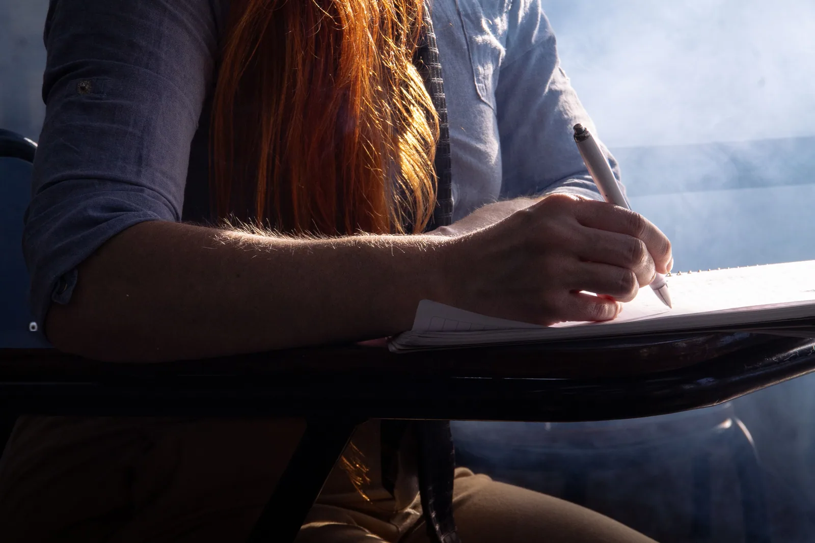 Estudante escreve em um caderno apoiado na carteira, em contraluz