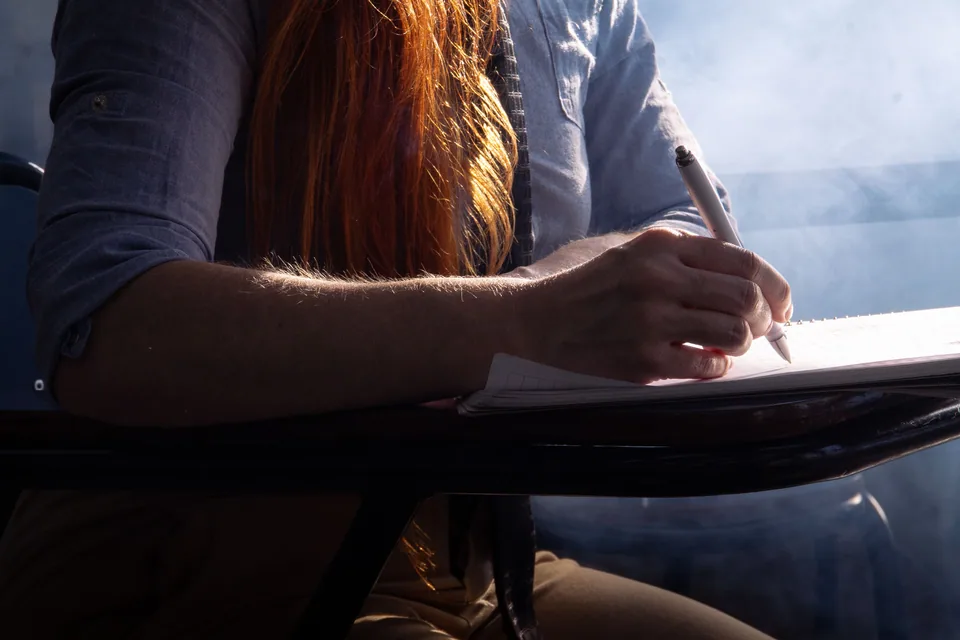 Estudante escreve em um caderno apoiado na carteira, em contraluz