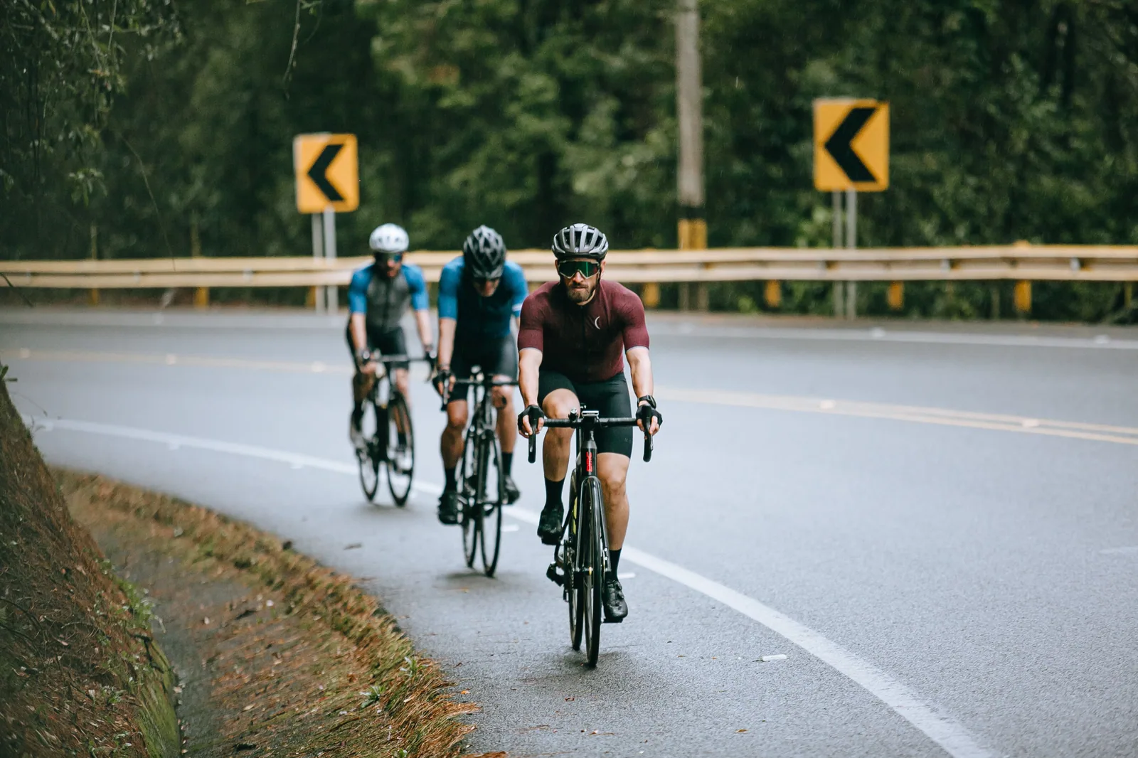 Três ciclistas de capacete pedalam em fila numa pista de asfalto cercada de árvores