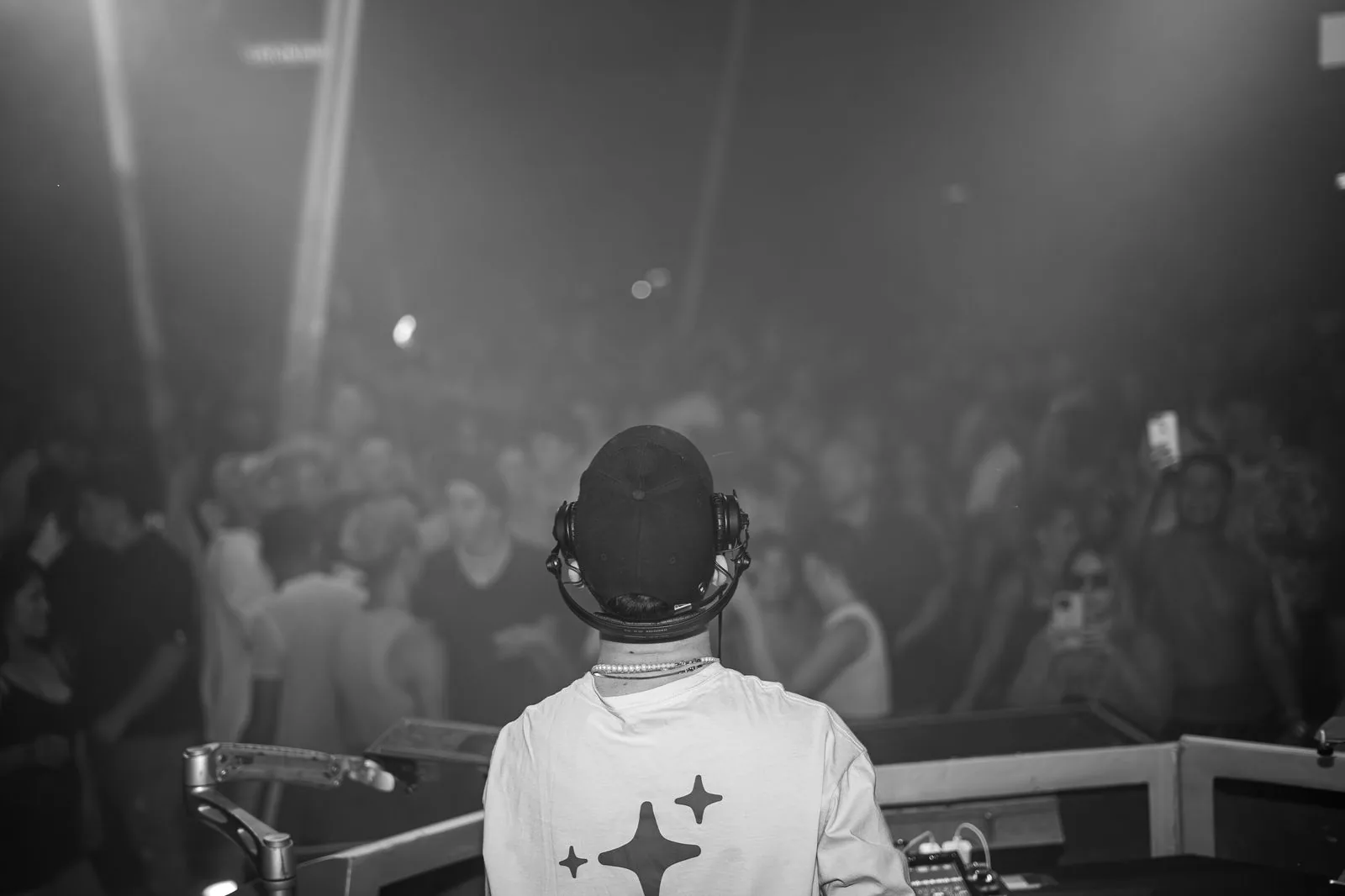 DJ de costas, de boné e fones, diante de uma pista cheia em preto e branco (foto ilustrativa de banco de imagens)