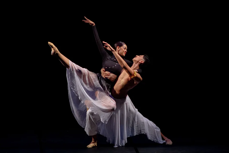 Dupla de bailarinos em pose de balé sobre palco com fundo preto, a bailarina de saia branca esvoaçante