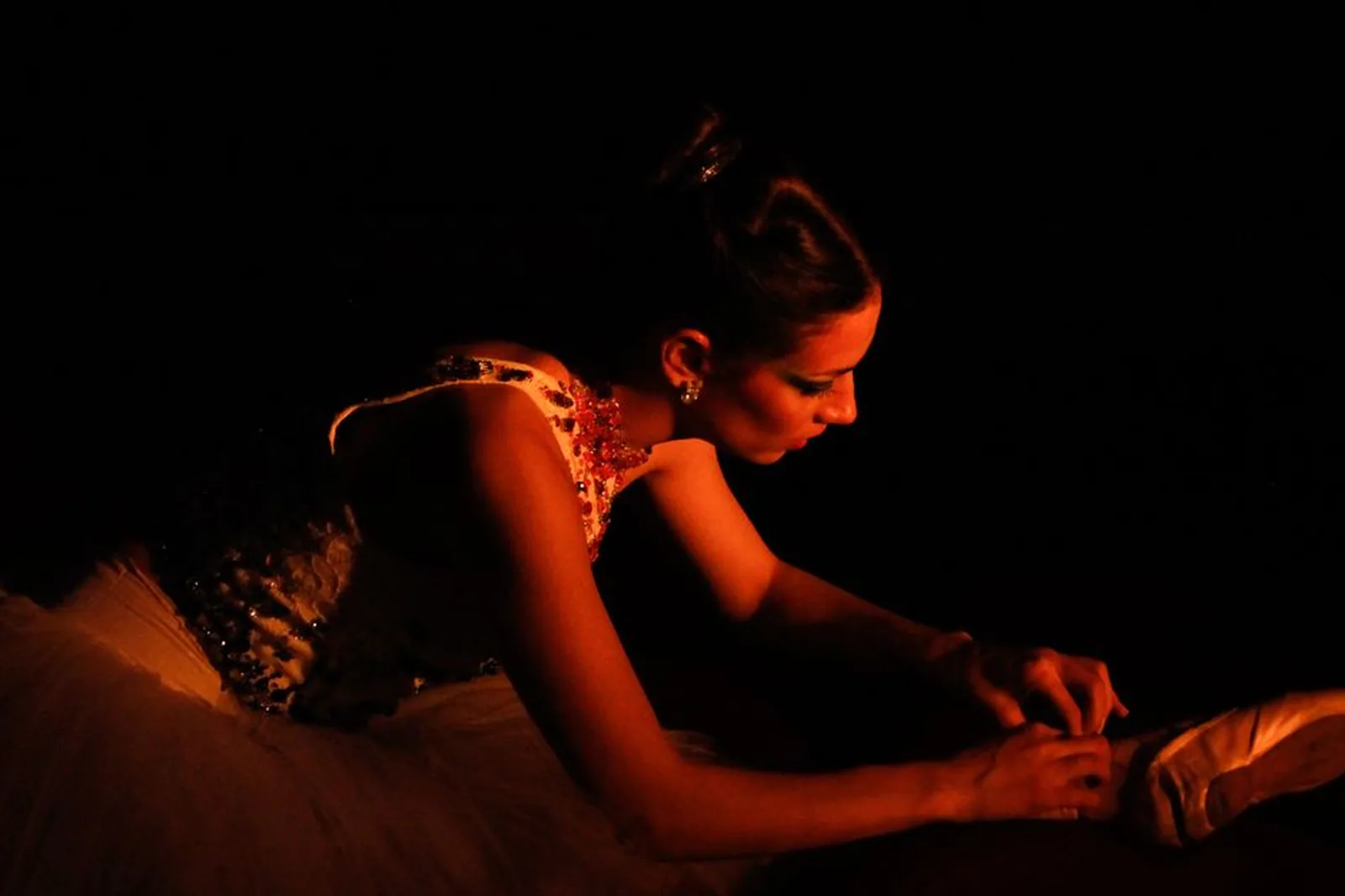 Bailarina de tutu ajusta a sapatilha de ponta em palco escuro, iluminada por luz quente
