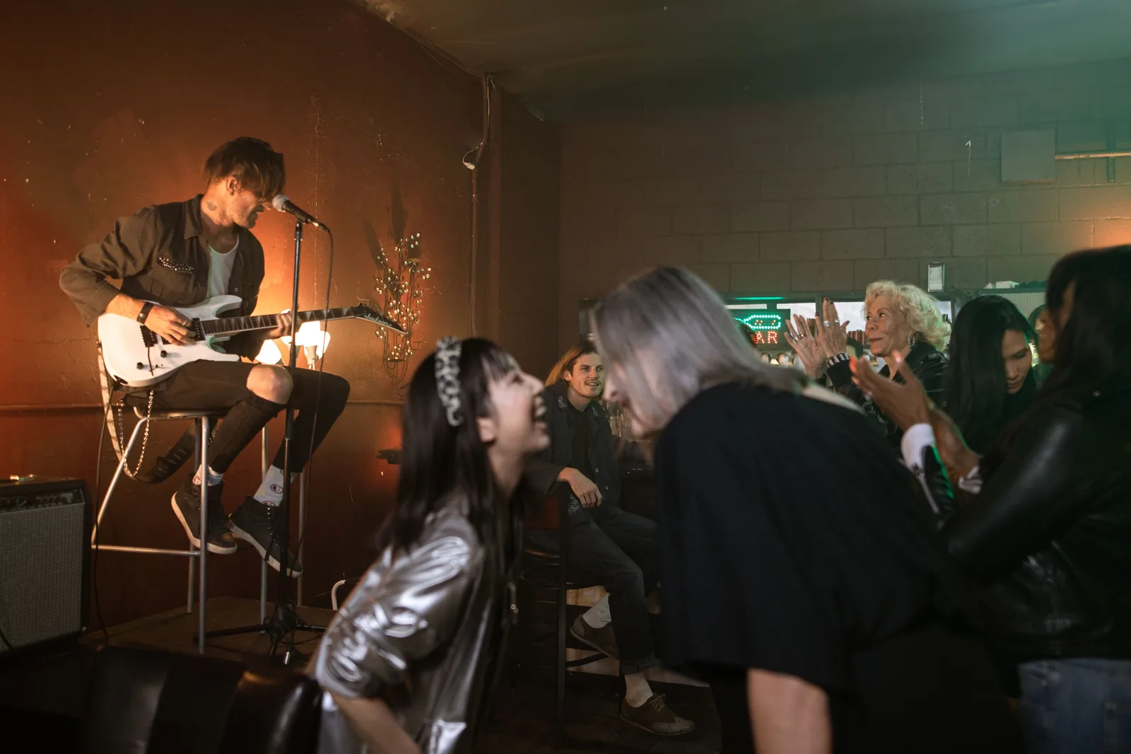 Músico tocando guitarra em palco pequeno de bar, com plateia próxima e luz quente ao fundo