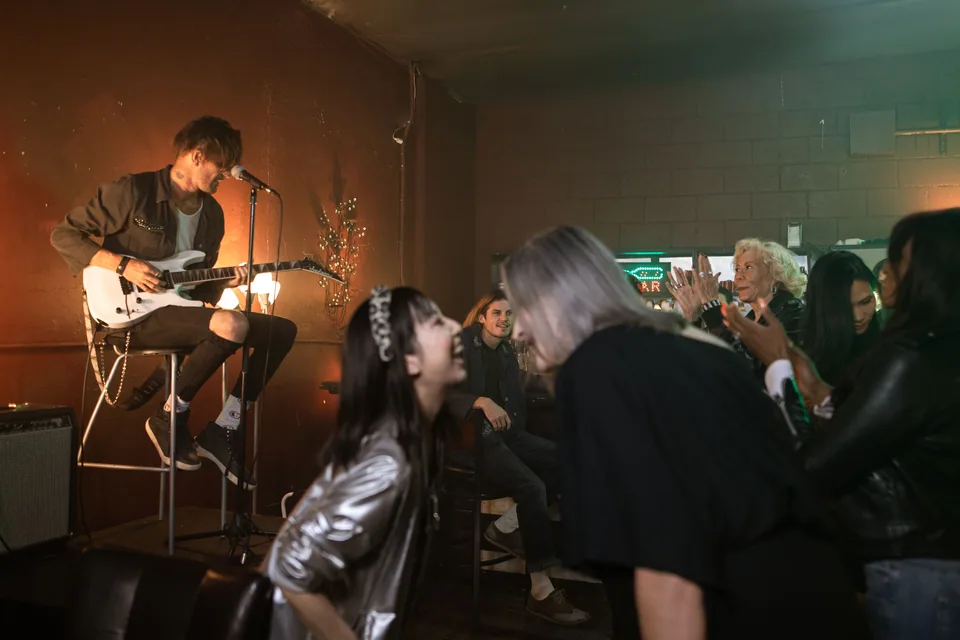 Músico tocando guitarra em palco pequeno de bar, com plateia próxima e luz quente ao fundo