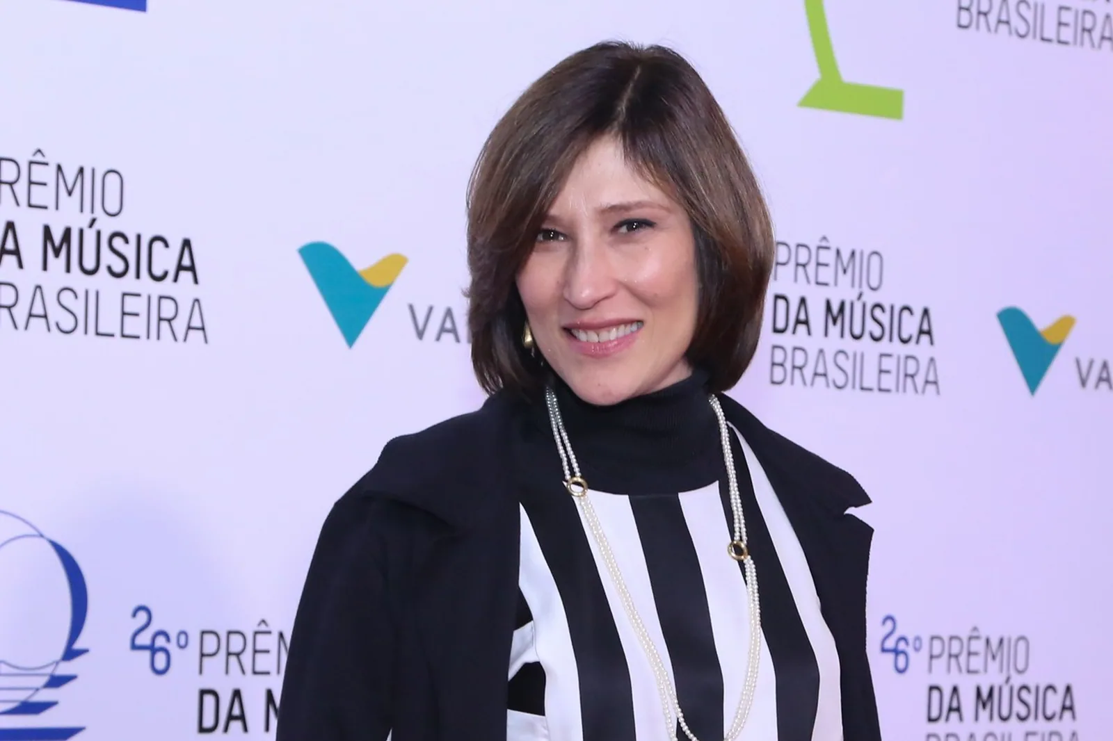 Beth Goulart sorrindo diante de painel de evento, de blusa listrada em preto e branco e casaco preto