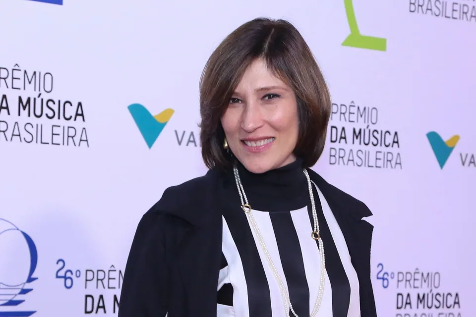 Beth Goulart sorrindo diante de painel de evento, de blusa listrada em preto e branco e casaco preto