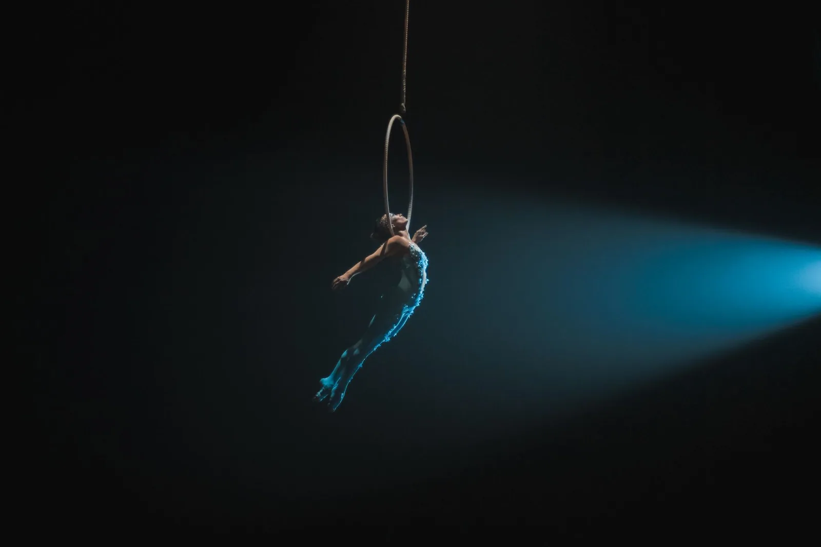 Artista de circo suspensa em um aro aéreo, iluminada por um facho de luz azul em palco escuro