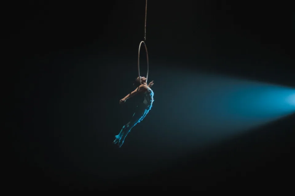 Artista de circo suspensa em um aro aéreo, iluminada por um facho de luz azul em palco escuro