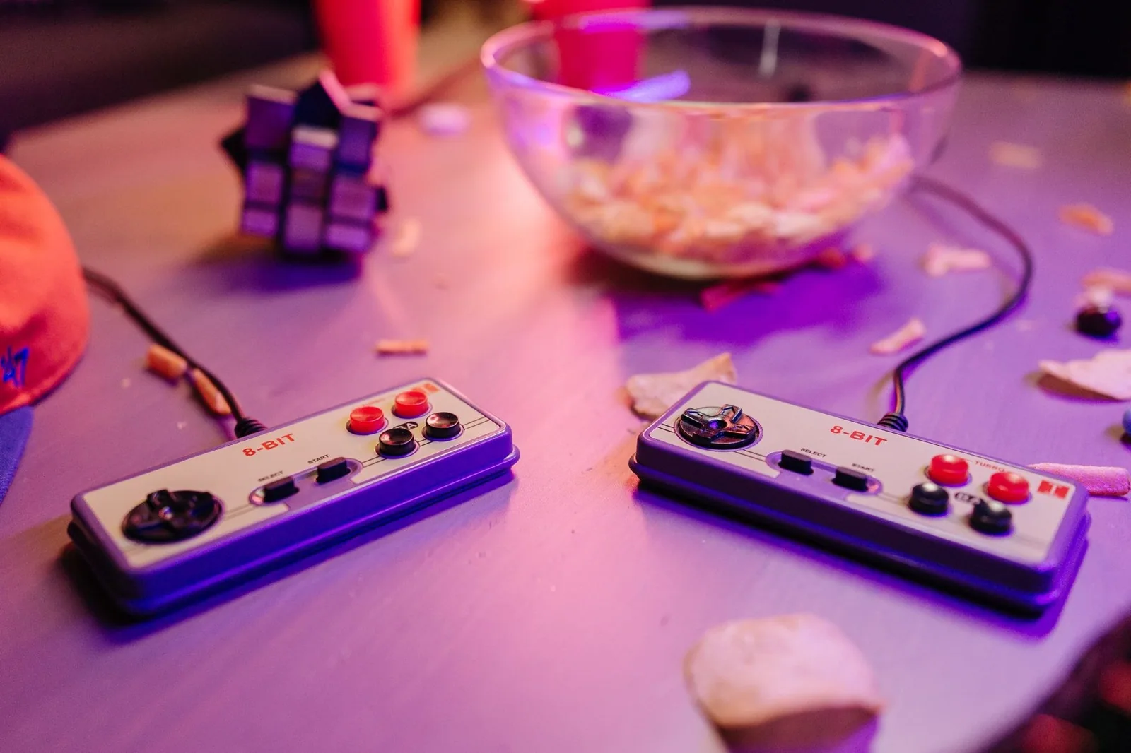 Dois controles de videogame 8 bits sobre uma mesa, com tigela de salgadinhos ao fundo