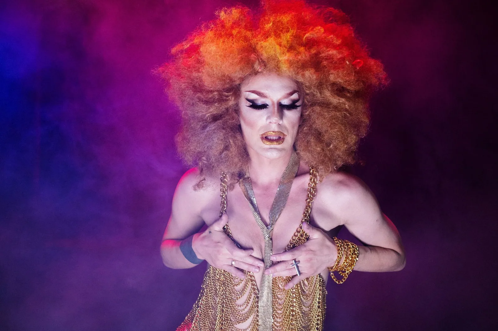 Artista drag de peruca laranja e figurino dourado sob luz roxa e fumaça