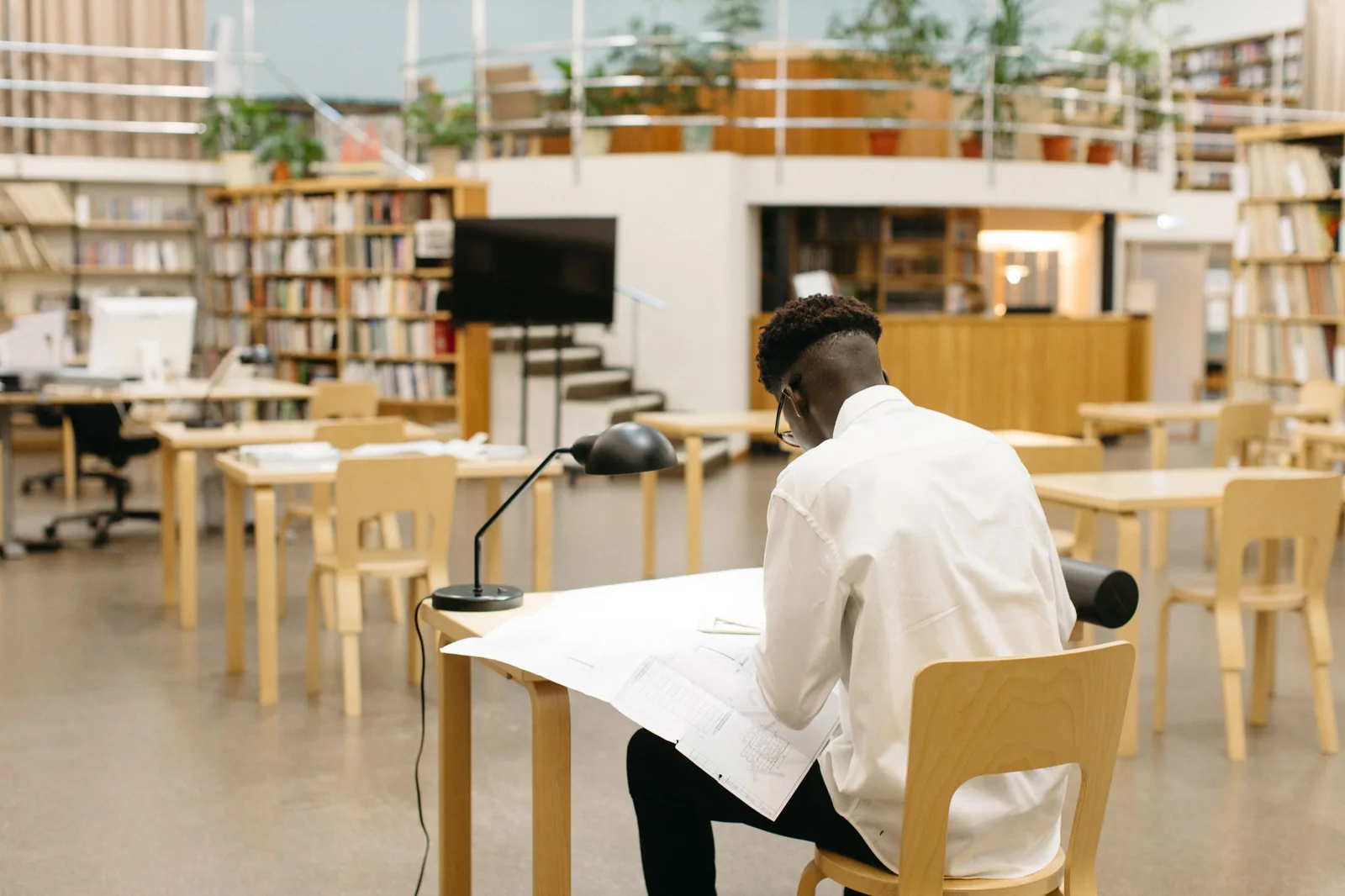 Estudante de camisa branca debruçado sobre papéis numa mesa de madeira, com luminária, em biblioteca ampla com estantes de livros ao fundo.