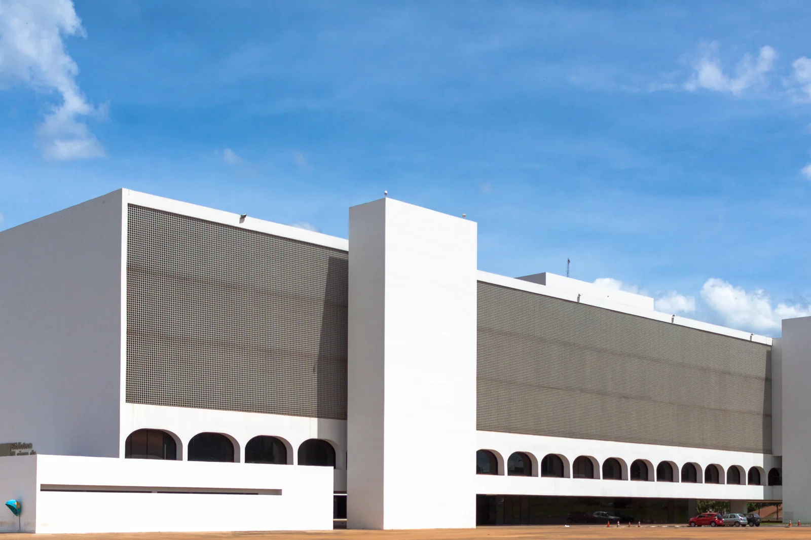 Fachada branca e modernista da Biblioteca Nacional de Brasília sob céu azul. Foto de arquivo do Wikimedia Commons.