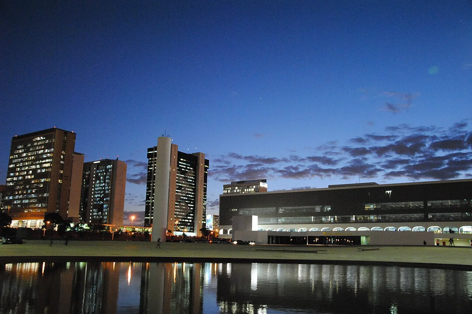 Prédio da Biblioteca Nacional de Brasília ao entardecer, visto do outro lado do espelho d'água, com edifícios do Setor Bancário ao fundo