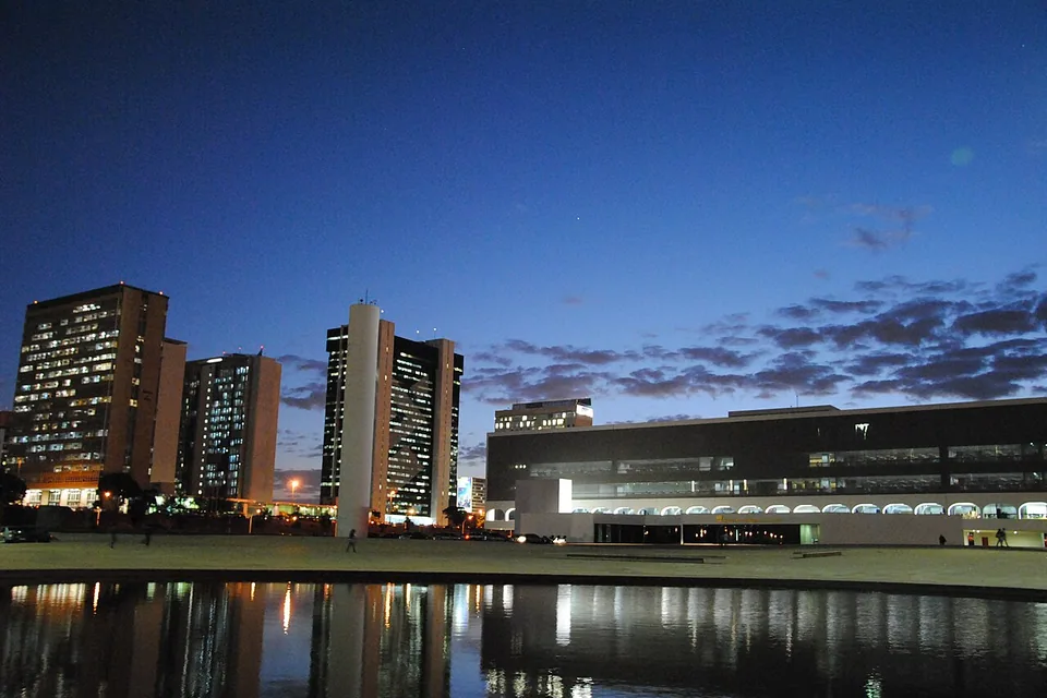 Prédio da Biblioteca Nacional de Brasília ao entardecer, visto do outro lado do espelho d'água, com edifícios do Setor Bancário ao fundo