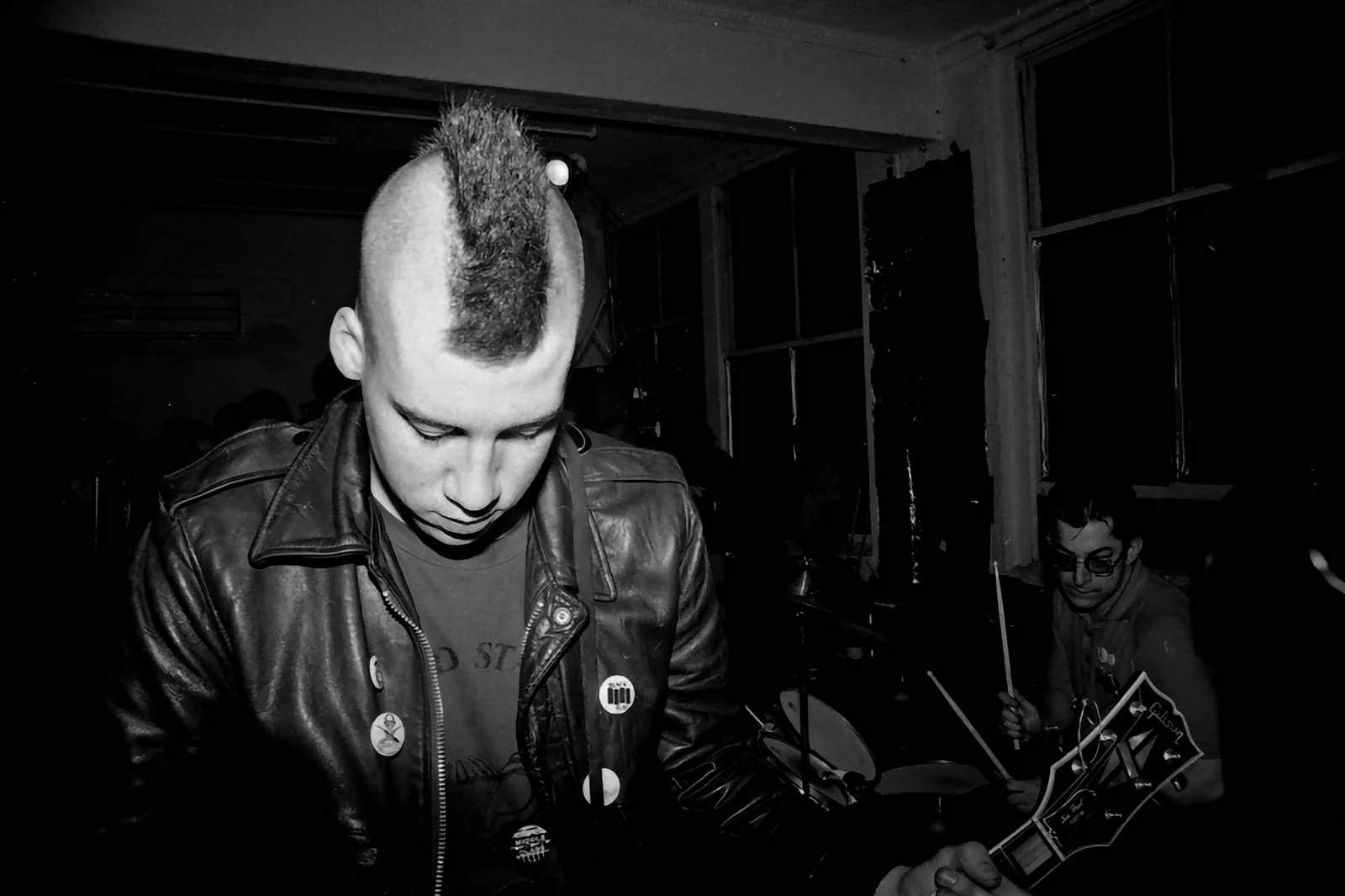 Músico de moicano e jaqueta de couro em show punk em preto e branco, com baterista ao fundo
