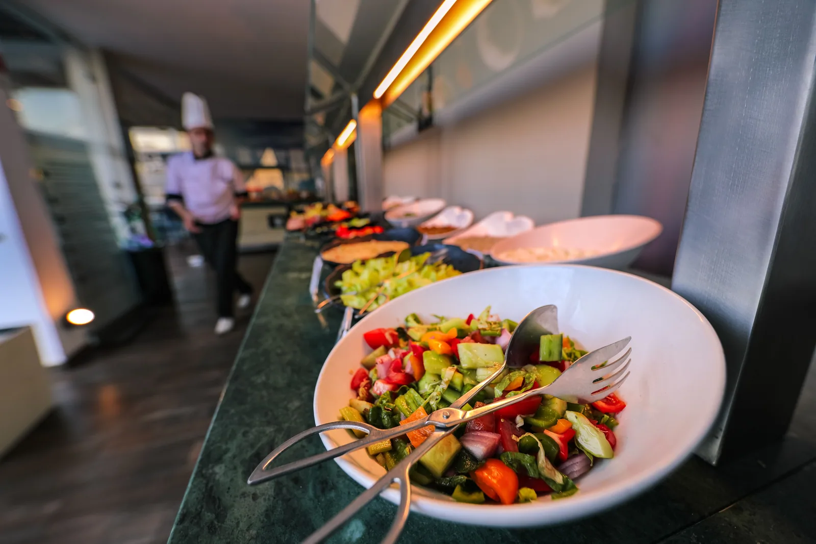 Travessa de salada colorida em balcão de buffet, com cozinheiro ao fundo