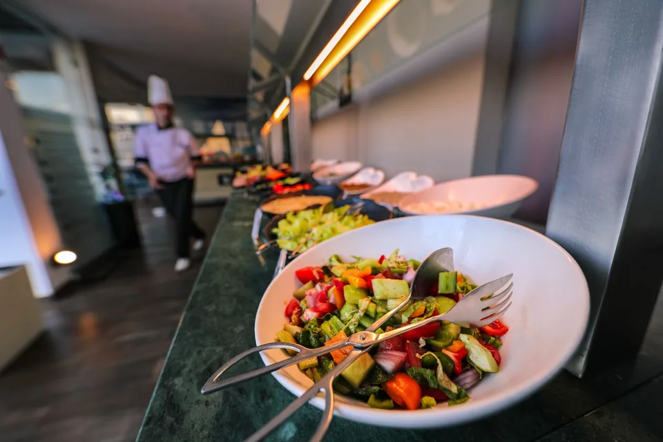 Travessa de salada colorida em balcão de buffet, com cozinheiro ao fundo
