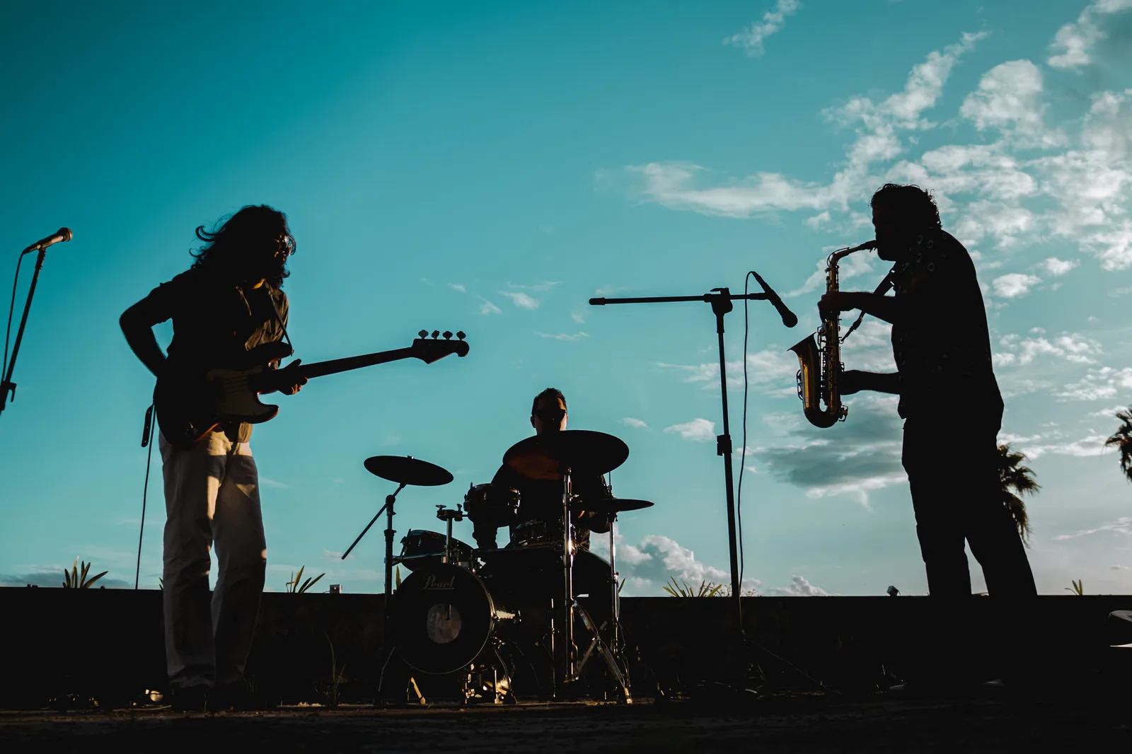 Silhueta de uma banda com baixo, bateria e saxofone tocando ao ar livre no fim da tarde