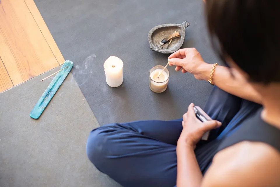 Pessoa sentada acendendo uma vela ao lado de incenso e de um pedaço de palo santo, sobre um tapete de meditação
