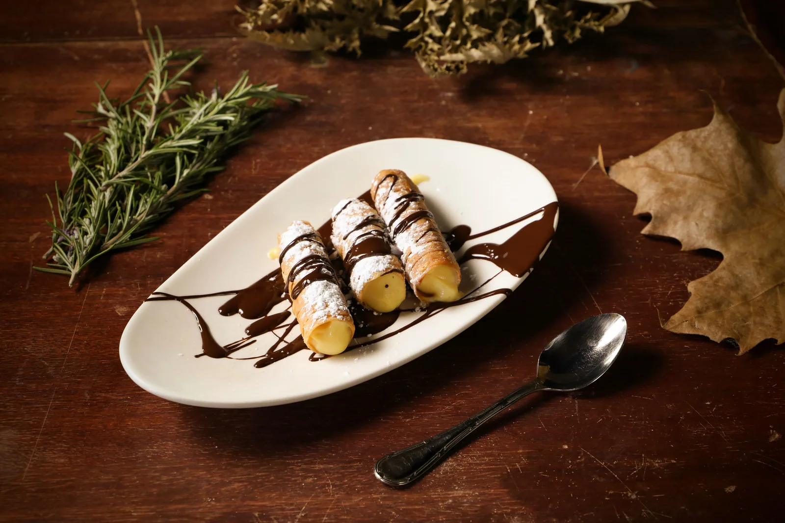 Três cannoli polvilhados com açúcar sobre prato branco oval, com calda de chocolate desenhada ao redor