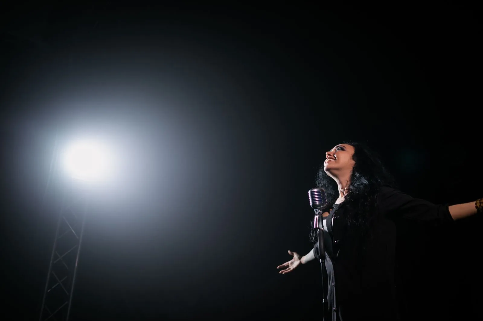 Cantora com os braços abertos diante de um microfone antigo e de um refletor, em palco escuro (foto ilustrativa)