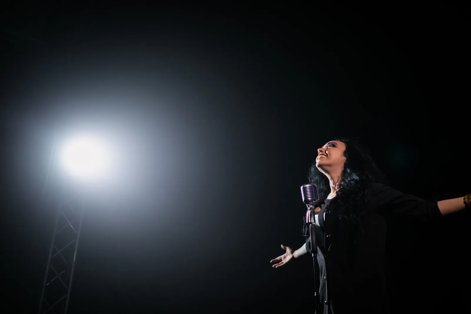 Cantora de braços abertos diante de microfone antigo em palco escuro iluminado por refletor