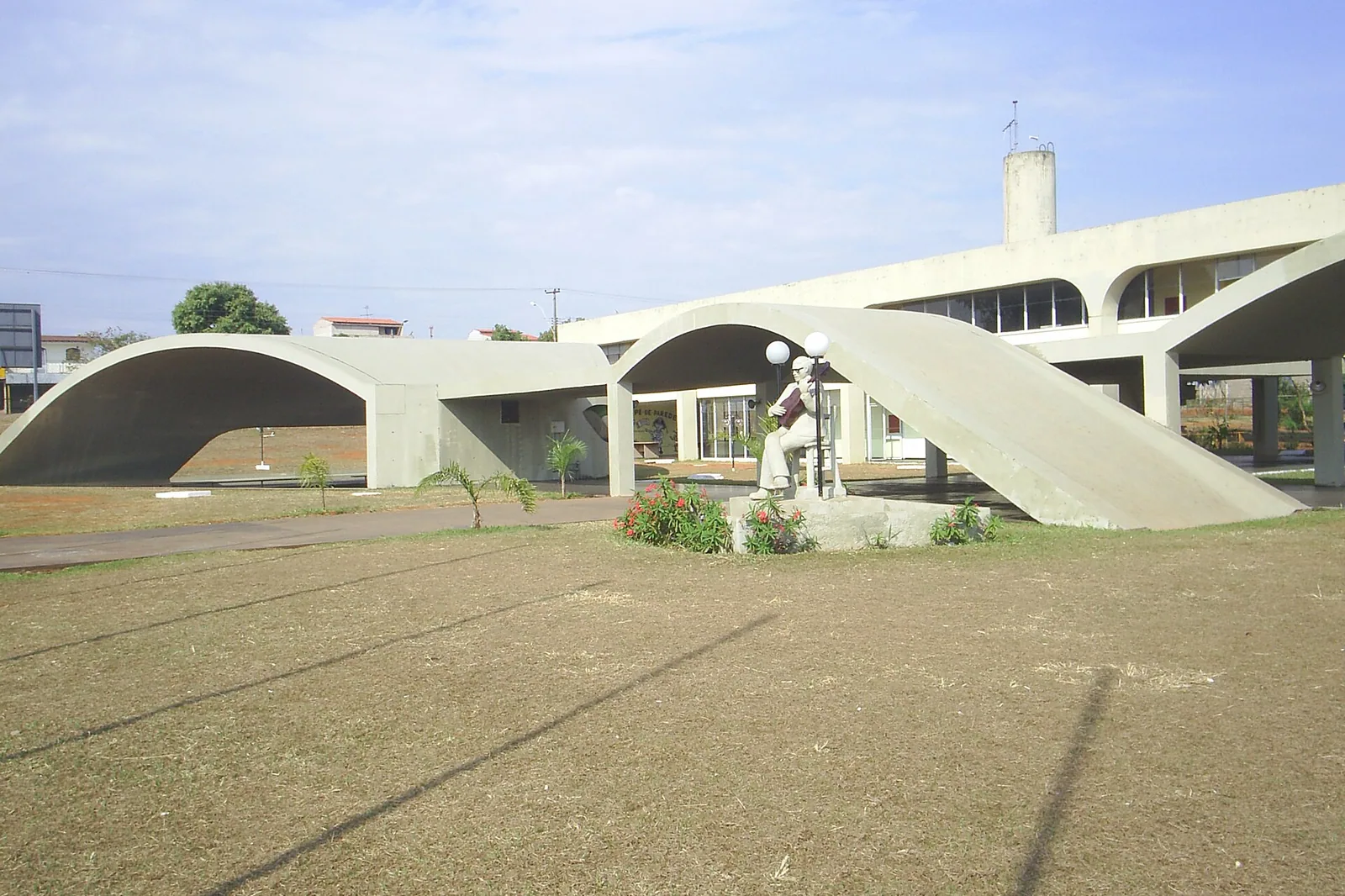 Prédio de concreto da Casa do Cantador, com arcos curvos que descem até o chão e estátua de um cantador com viola no gramado à frente.