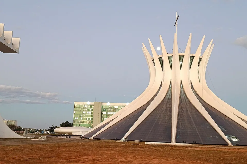 Catedral Metropolitana de Brasília vista da Esplanada no fim de tarde, com o campanário à esquerda e a lua cheia acima das colunas brancas