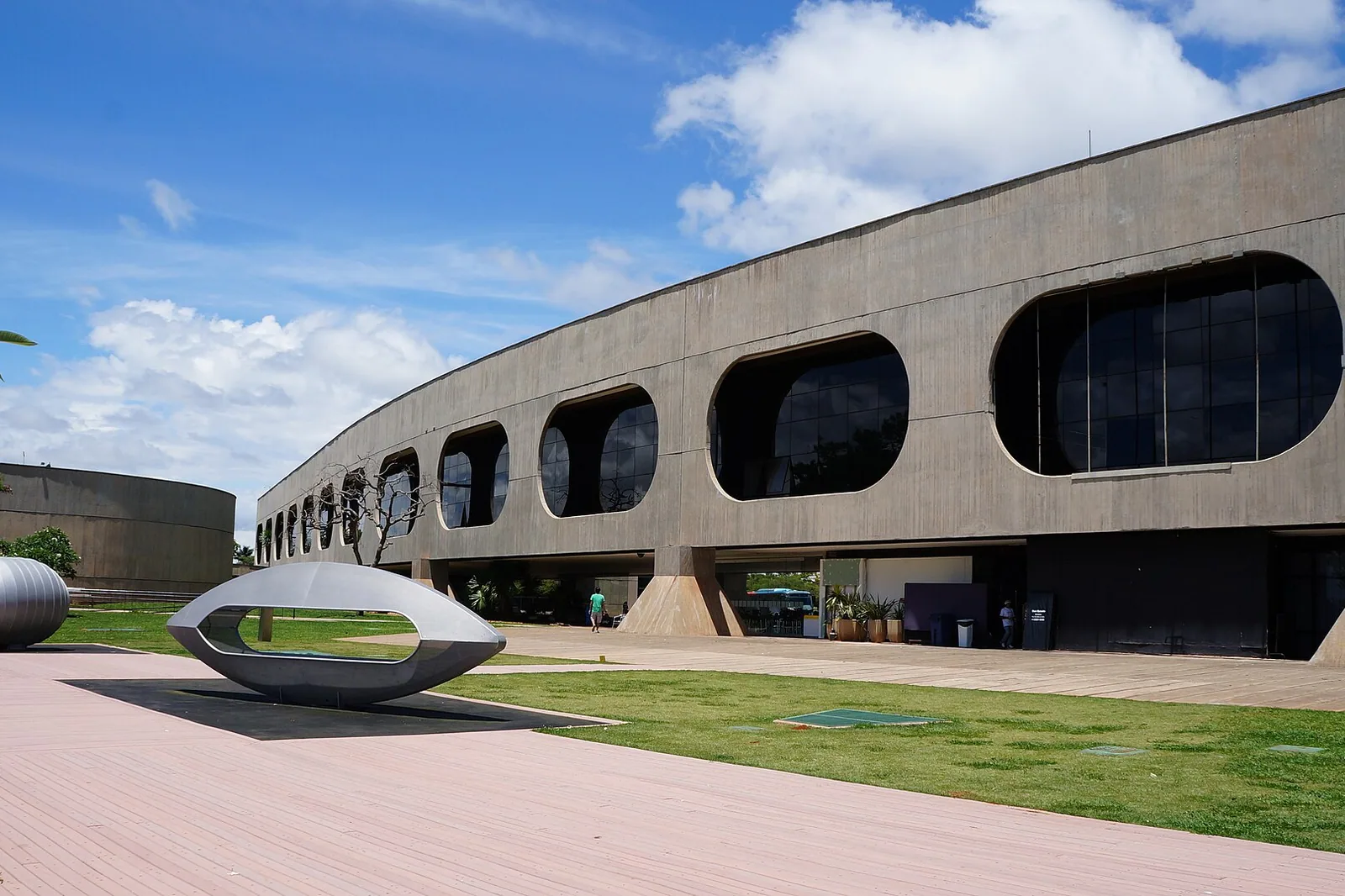 Fachada curva de concreto do CCBB Brasília com janelas ovais, gramado e esculturas metálicas em primeiro plano