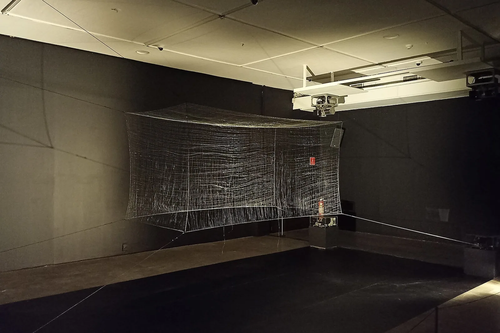 Sala escura de exposição no CCBB Brasília, com uma grande instalação de fios suspensa no centro e holofote ao fundo