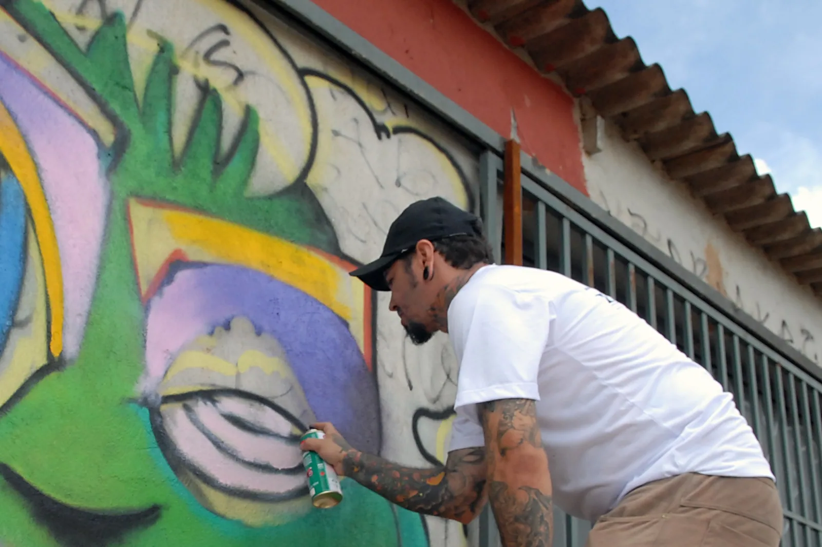 Grafiteiro de boné e camiseta branca pinta um muro colorido com spray em Ceilândia