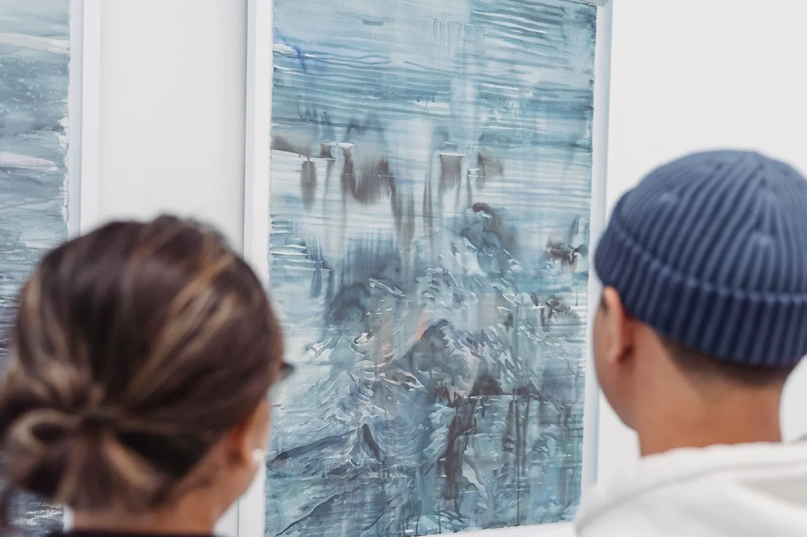 Duas pessoas de costas olham uma pintura em aquarela azulada emoldurada na parede branca de uma galeria (foto ilustrativa)