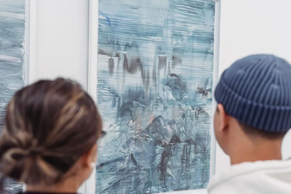 Duas pessoas de costas olham uma pintura em aquarela azulada emoldurada na parede branca de uma galeria (foto ilustrativa)