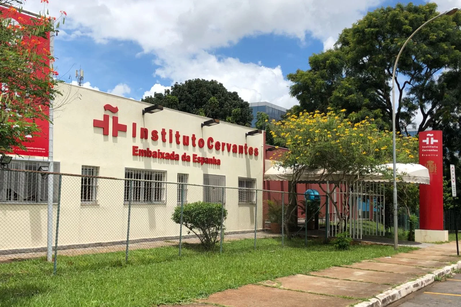 Fachada do Instituto Cervantes de Brasília, prédio claro com letreiro vermelho, árvores floridas e totem vermelho na entrada