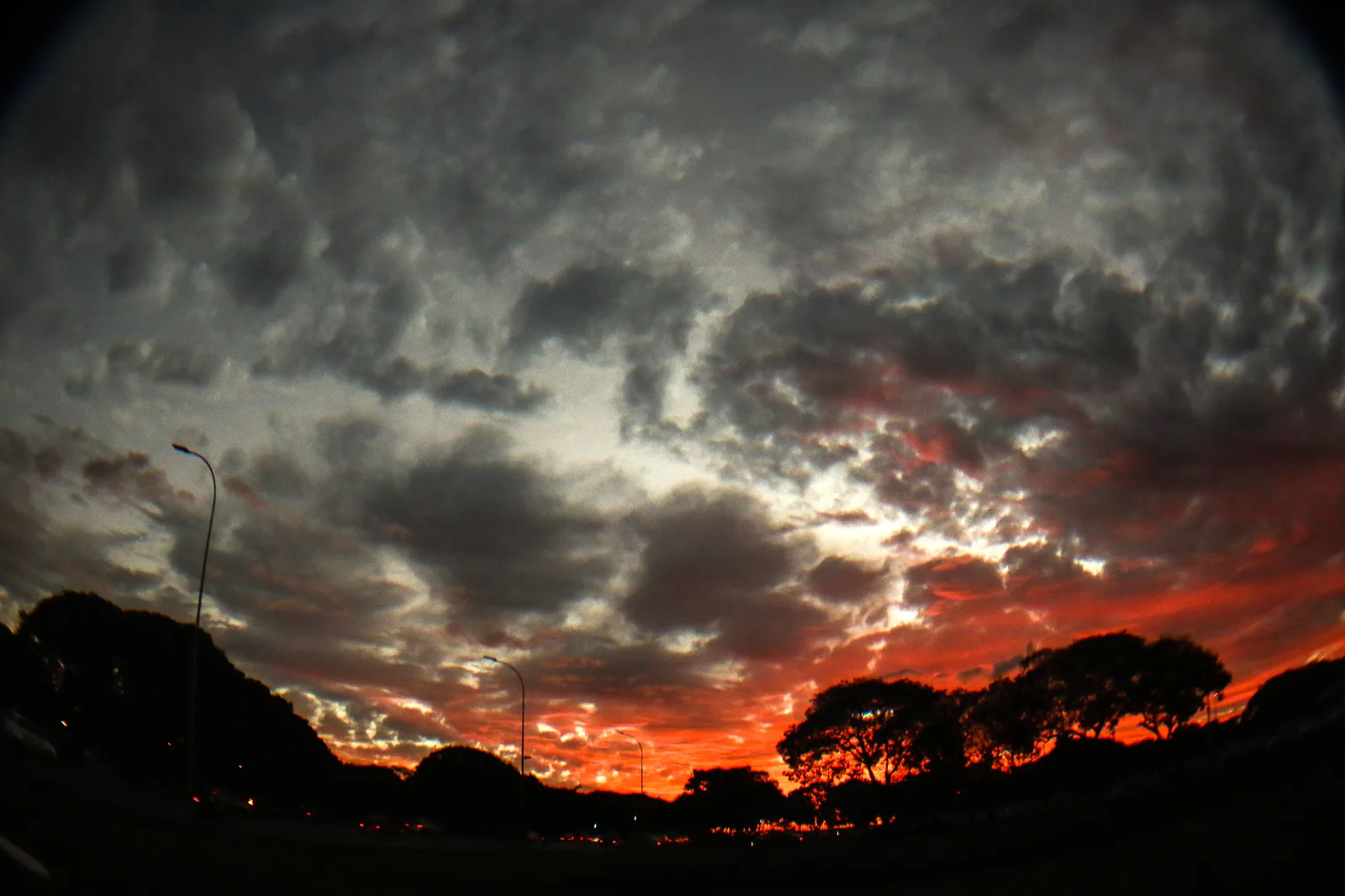 Céu de Brasília no fim do dia, com nuvens e faixa laranja do pôr do sol sobre silhuetas de árvores