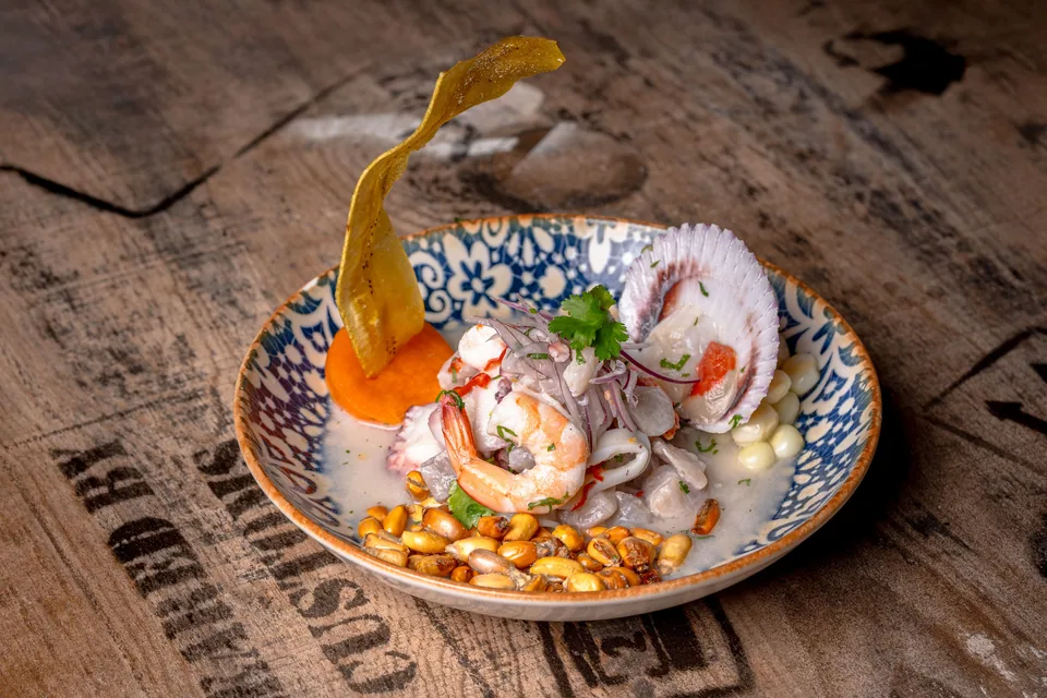 Ceviche com camarão, vieira, milho cancha e chip de banana servido em prato azul sobre mesa de madeira