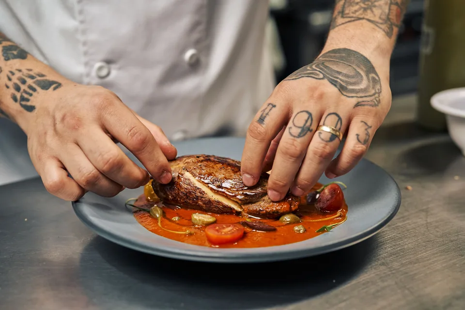 Mãos tatuadas de um cozinheiro de jaleco finalizam um prato de carne com molho de tomate, alcaparras e tomate-cereja, em foto de arquivo.