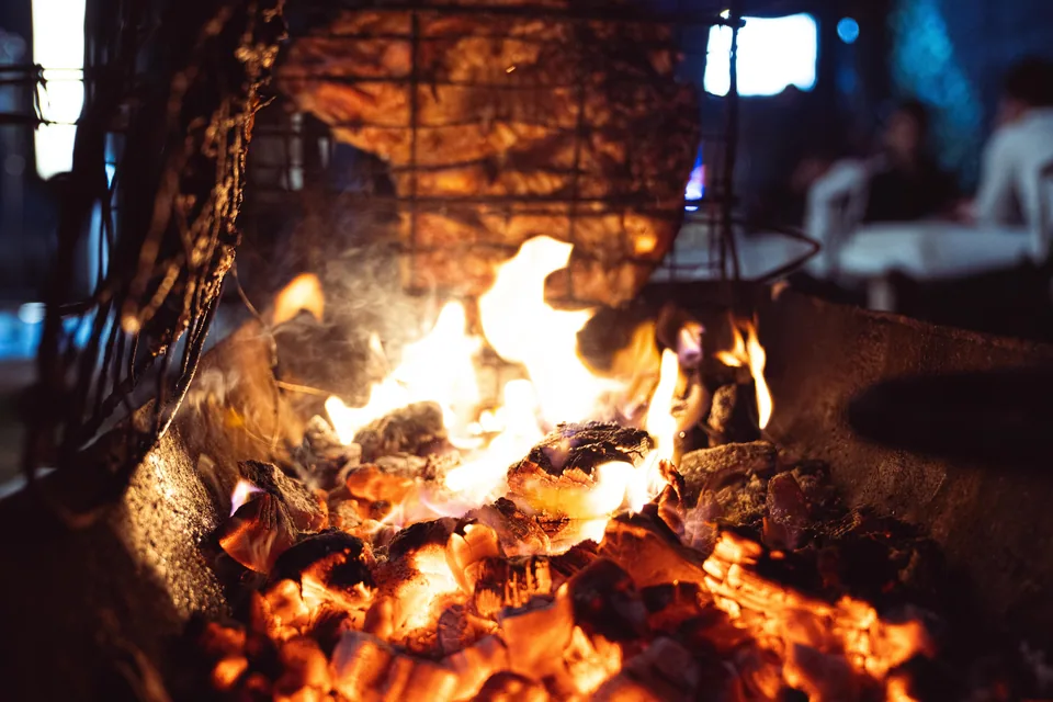 Carne assando em grelha sobre brasas acesas em churrasqueira ao ar livre, com mesas ao fundo