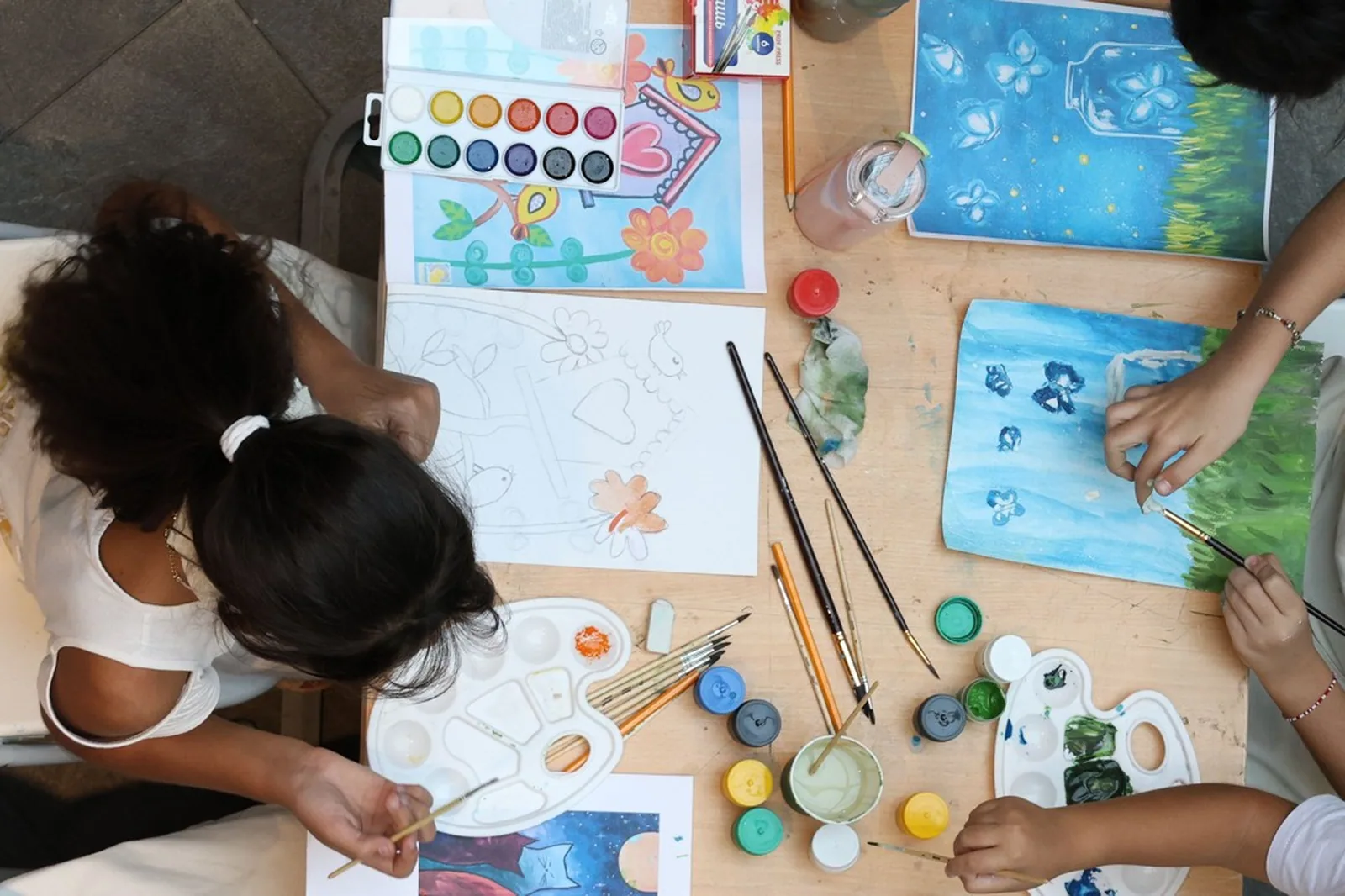 Crianças pintando com aquarela e guache em uma mesa de oficina, vistas de cima