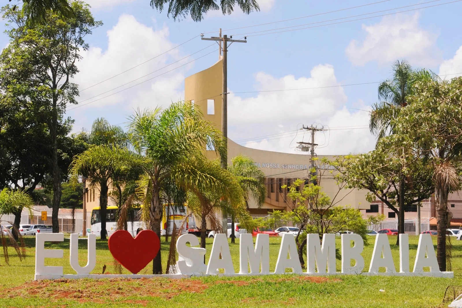 Letreiro branco com a frase Eu amo Samambaia em gramado, com palmeiras e uma igreja ao fundo
