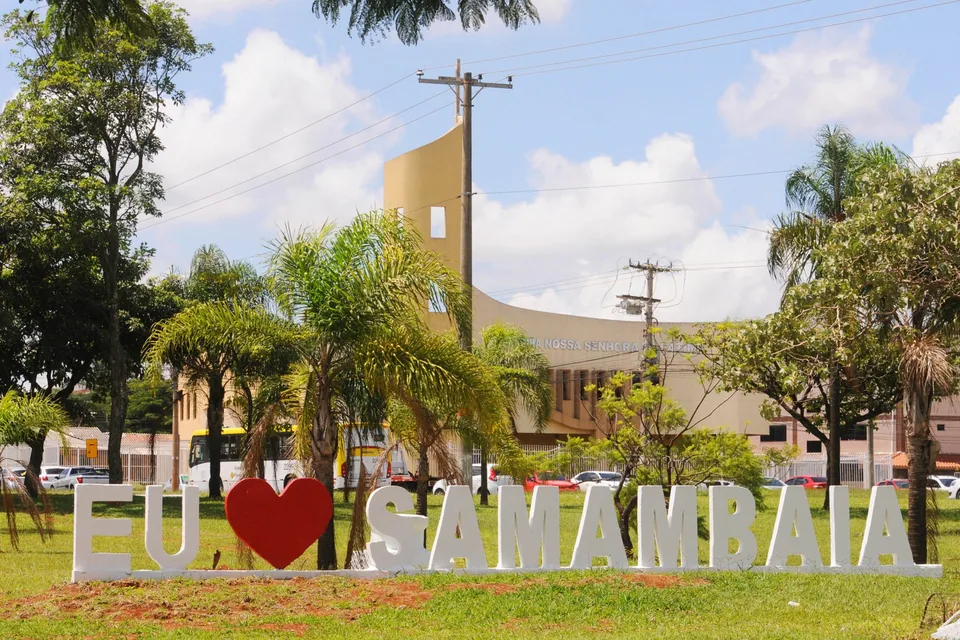 Letreiro branco com a frase Eu amo Samambaia em gramado, com palmeiras e uma igreja ao fundo