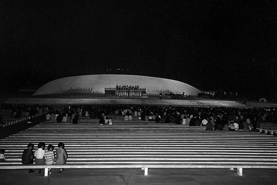 Foto antiga em preto e branco da Concha Acústica à noite, com coro no palco sob a concha e público sentado nos bancos de concreto da arquibancada.